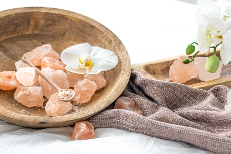 Komposisi spa dengan garam laut merah muda, bunga anggrek, handuk, dan batu pijat di atas meja kayu.